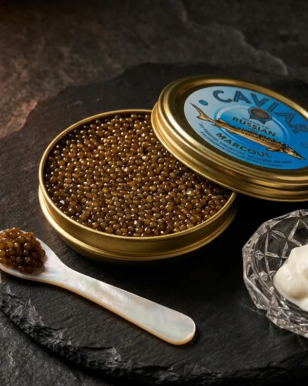 An open tin of Ossetra caviar with amber-to-charcoal pearls, a mother-of-pearl spoon and crème fraîche on dark slate.
